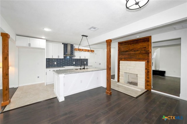 kitchen featuring white cabinetry, decorative light fixtures, an island with sink, light stone countertops, and wall chimney range hood