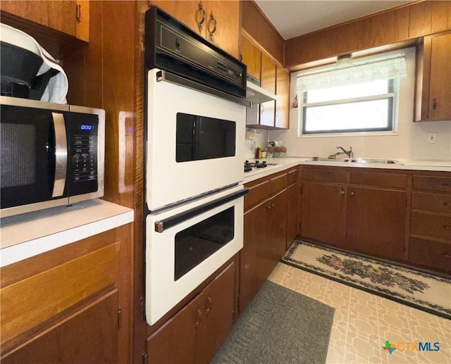 kitchen featuring under cabinet range hood, light countertops, brown cabinetry, white appliances, and a sink