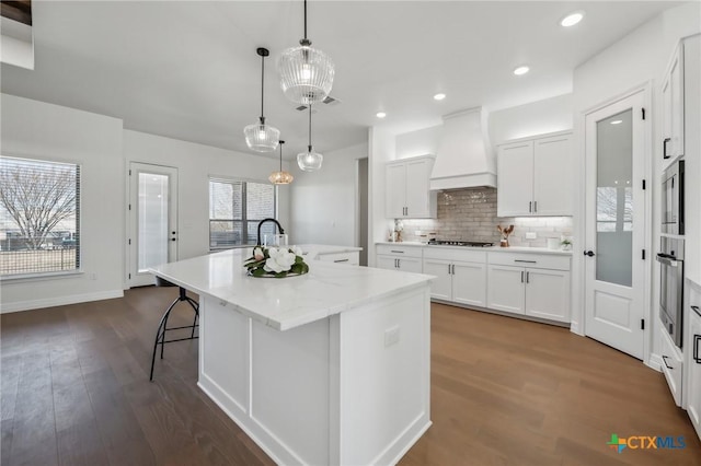 kitchen with pendant lighting, white cabinetry, an island with sink, backsplash, and custom range hood