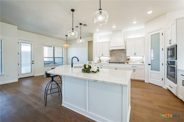 kitchen featuring white cabinetry, hanging light fixtures, an island with sink, custom range hood, and oven