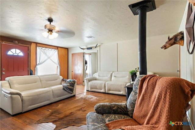 living room with ceiling fan, a textured ceiling, wood finished floors, visible vents, and a wood stove