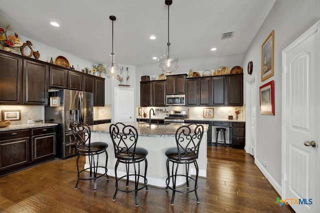 kitchen featuring a kitchen island with sink, dark wood-type flooring, hanging light fixtures, and appliances with stainless steel finishes