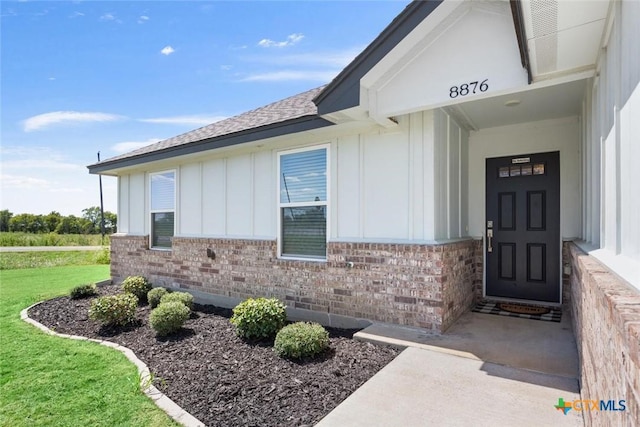 view of exterior entry with a shingled roof, a lawn, board and batten siding, and brick siding