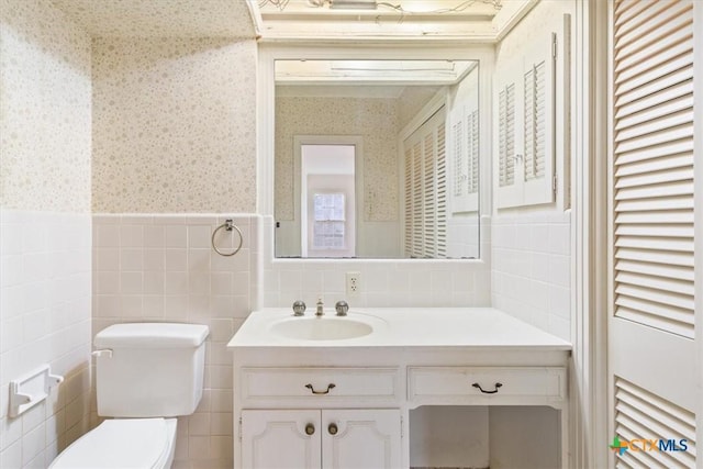 bathroom featuring toilet, a wainscoted wall, tile walls, vanity, and wallpapered walls