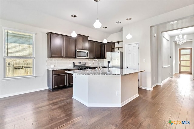 kitchen with appliances with stainless steel finishes, dark brown cabinets, light stone counters, a center island with sink, and decorative light fixtures