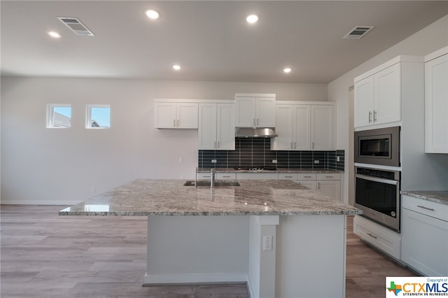 kitchen with white cabinetry, appliances with stainless steel finishes, light wood-type flooring, light stone countertops, and an island with sink