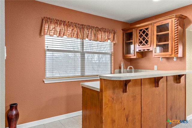 kitchen with a breakfast bar area, a peninsula, light countertops, brown cabinetry, and glass insert cabinets