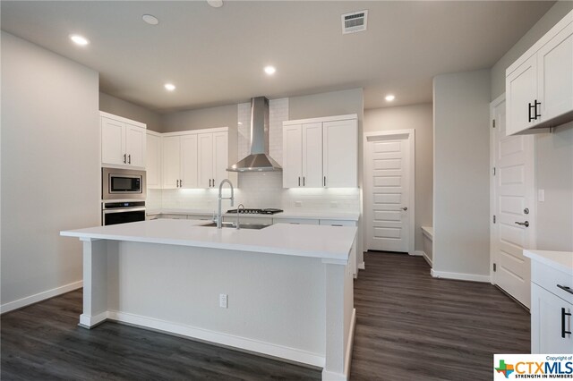 kitchen with wall chimney range hood, appliances with stainless steel finishes, sink, an island with sink, and dark wood-type flooring