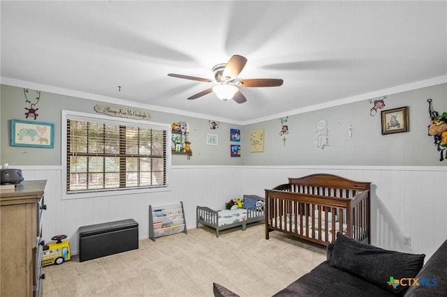 bedroom featuring light carpet, ceiling fan, a crib, and wainscoting