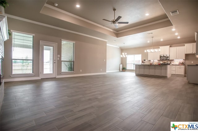 unfurnished living room featuring a wealth of natural light, ceiling fan with notable chandelier, dark hardwood / wood-style flooring, and a tray ceiling