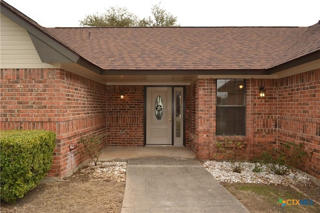 view of exterior entry featuring a shingled roof and brick siding