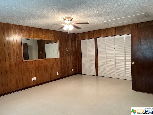 unfurnished bedroom featuring a textured ceiling, wood walls, ceiling fan, and two closets