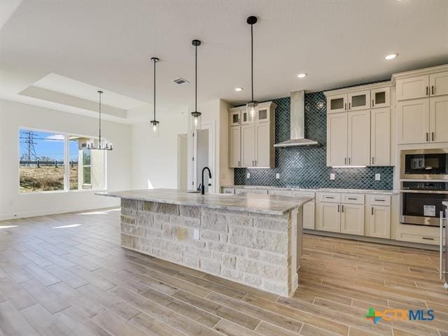 kitchen with wall chimney exhaust hood, light hardwood / wood-style flooring, oven, pendant lighting, and a kitchen island with sink