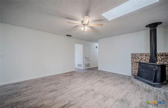 unfurnished living room with visible vents, a skylight, a wood stove, wood finished floors, and a ceiling fan