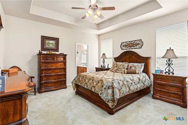 bedroom with a tray ceiling, light carpet, ensuite bathroom, and a ceiling fan