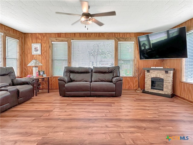 living room featuring wood walls, ceiling fan, a textured ceiling, a stone fireplace, and light wood-type flooring