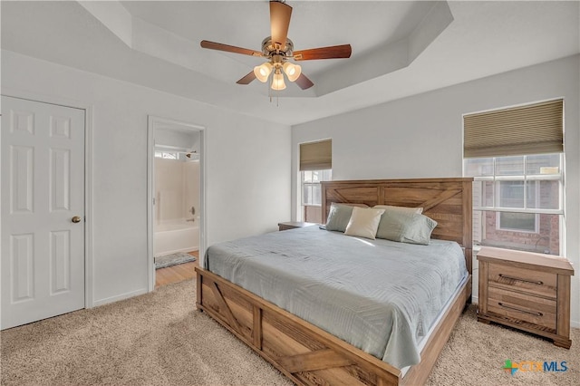 bedroom featuring ensuite bathroom, ceiling fan, a tray ceiling, and light colored carpet