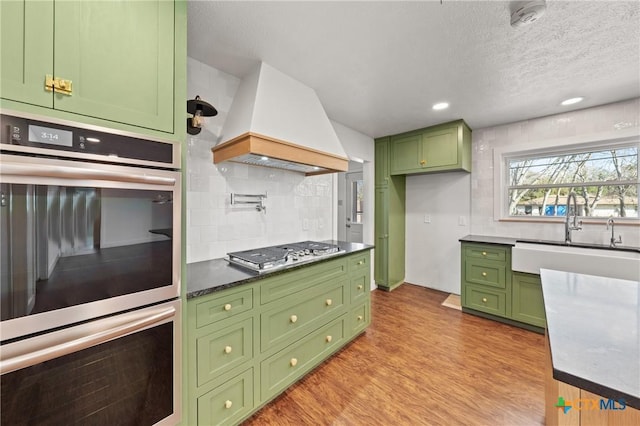 kitchen with a sink, light wood-style floors, appliances with stainless steel finishes, custom exhaust hood, and green cabinetry