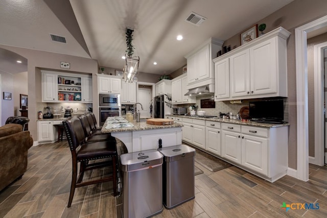 kitchen featuring appliances with stainless steel finishes, range hood, white cabinets, a kitchen island with sink, and light stone countertops