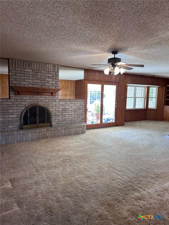 unfurnished living room featuring a textured ceiling, carpet floors, wooden walls, a fireplace, and ceiling fan