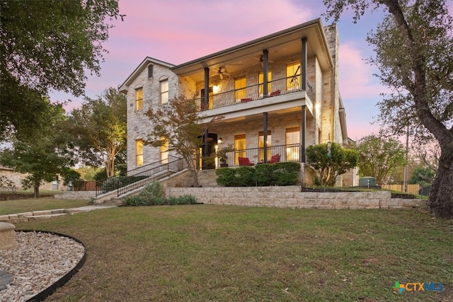 back house at dusk featuring a lawn, ceiling fan, and a balcony