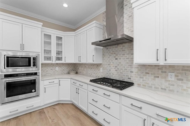 kitchen with appliances with stainless steel finishes, white cabinetry, ornamental molding, light stone countertops, and wall chimney exhaust hood