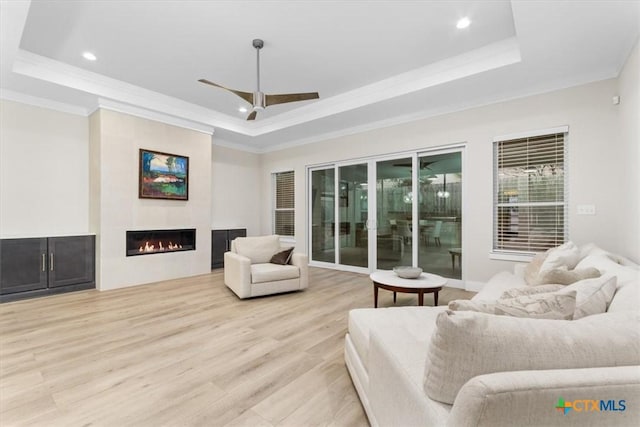 living area featuring a glass covered fireplace, a raised ceiling, light wood-style floors, and ornamental molding