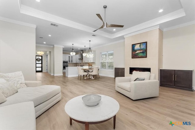 living room featuring crown molding, a tray ceiling, a fireplace, ceiling fan with notable chandelier, and light wood-type flooring