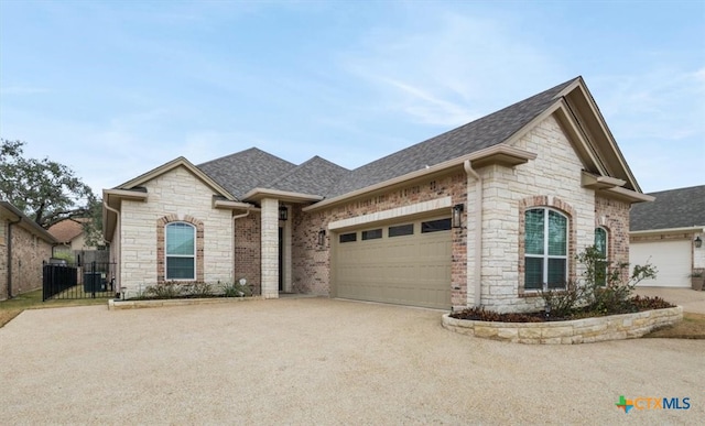 french country inspired facade featuring a garage, roof with shingles, concrete driveway, and fence