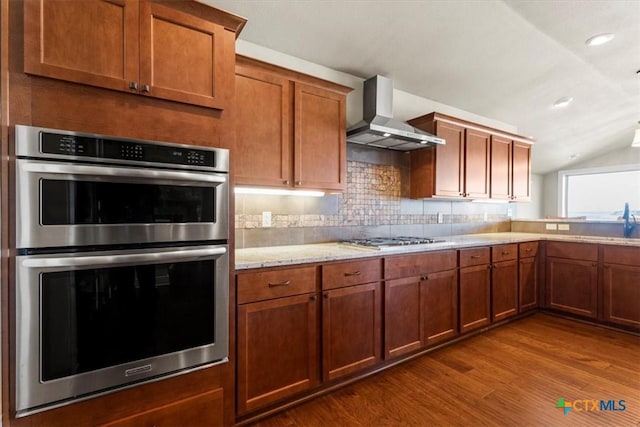 kitchen featuring dark wood-type flooring, vaulted ceiling, stainless steel appliances, decorative backsplash, and wall chimney range hood