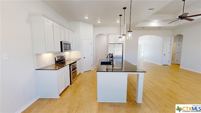 kitchen with light wood-type flooring, an island with sink, decorative light fixtures, white cabinetry, and stainless steel appliances