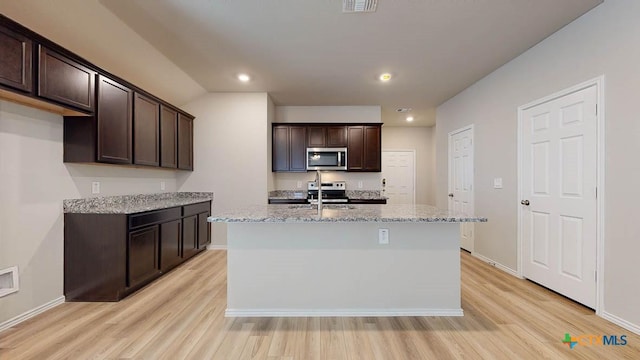 kitchen with light stone counters, dark brown cabinets, stainless steel appliances, a kitchen island with sink, and light hardwood / wood-style flooring