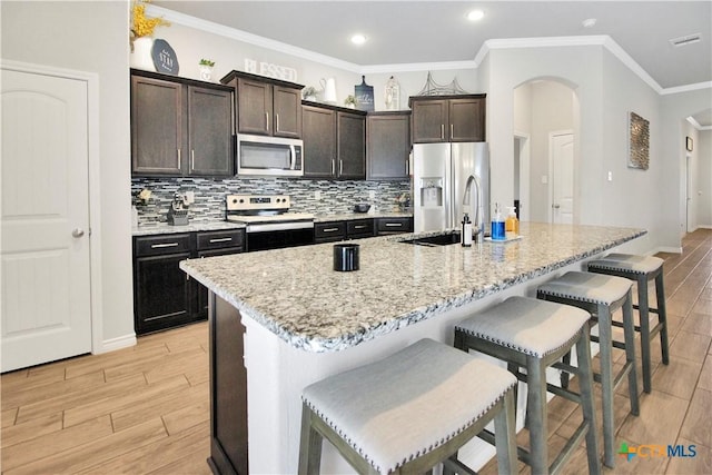 kitchen featuring appliances with stainless steel finishes, a breakfast bar area, backsplash, a kitchen island with sink, and dark brown cabinets