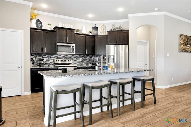 kitchen featuring stainless steel appliances, an island with sink, light stone counters, and dark brown cabinetry