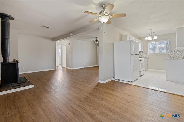 unfurnished living room featuring visible vents, light wood finished floors, baseboards, a wood stove, and ceiling fan with notable chandelier