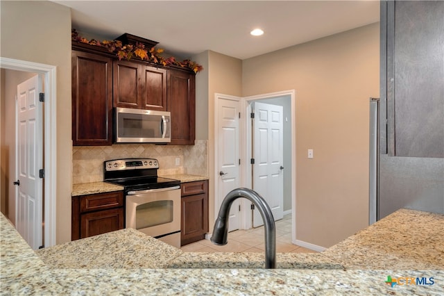 kitchen with light tile patterned floors, appliances with stainless steel finishes, backsplash, dark brown cabinetry, and light stone countertops