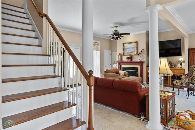 living area featuring a glass covered fireplace, crown molding, decorative columns, ceiling fan, and stairs