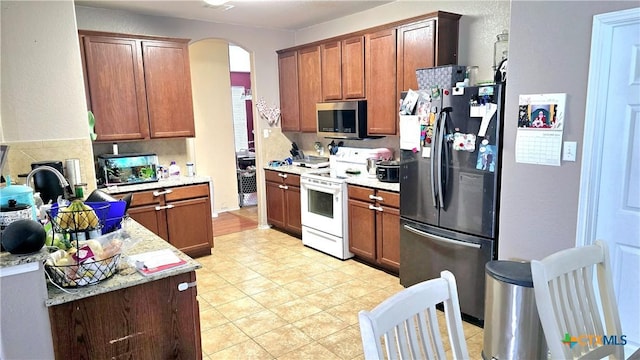 kitchen featuring arched walkways, light tile patterned floors, light stone counters, stainless steel appliances, and brown cabinetry