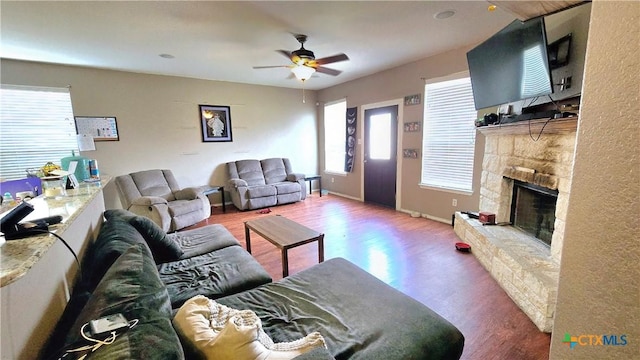 living room featuring ceiling fan, a stone fireplace, wood finished floors, and baseboards