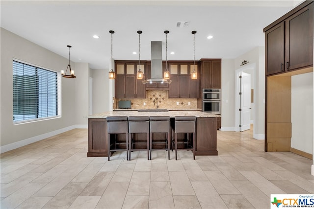 kitchen with exhaust hood, backsplash, an island with sink, and stainless steel double oven