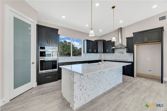 kitchen with a center island with sink, hanging light fixtures, wall chimney range hood, double oven, and light hardwood / wood-style flooring