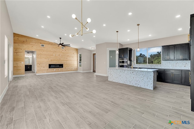 kitchen with stainless steel double oven, a kitchen island with sink, wood walls, hanging light fixtures, and light hardwood / wood-style flooring