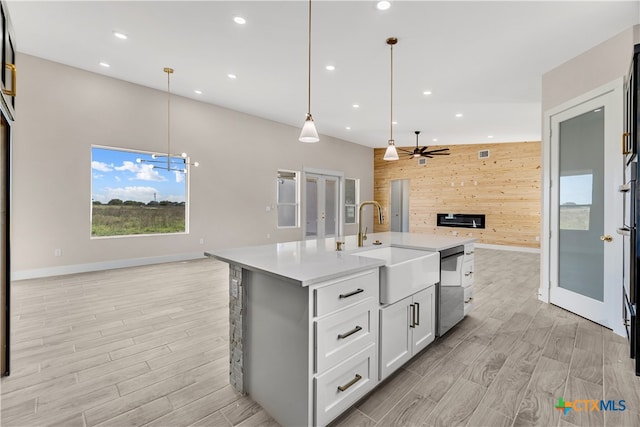 kitchen featuring dishwasher, wooden walls, sink, a kitchen island with sink, and pendant lighting