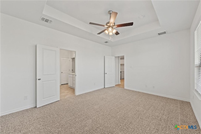 unfurnished bedroom with a tray ceiling, ensuite bath, ceiling fan, and light carpet