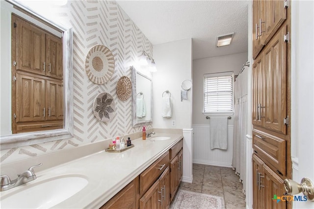 bathroom with a textured ceiling, wainscoting, a sink, and wallpapered walls