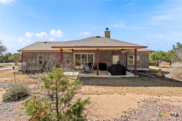 back of house with a shingled roof, brick siding, a patio, and a chimney