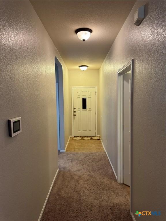 hallway featuring arched walkways, a textured wall, light carpet, a textured ceiling, and baseboards