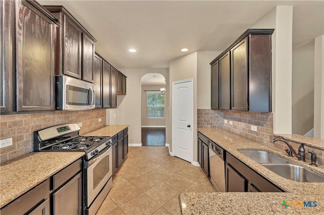 kitchen with a sink, arched walkways, appliances with stainless steel finishes, light stone countertops, and dark brown cabinets