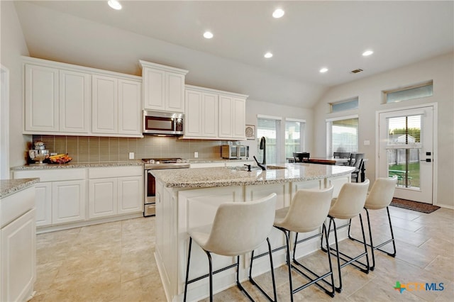 kitchen with stainless steel appliances, vaulted ceiling, a kitchen island with sink, white cabinets, and decorative backsplash