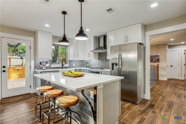 kitchen featuring wall chimney exhaust hood, stainless steel appliances, a kitchen island, pendant lighting, and white cabinets